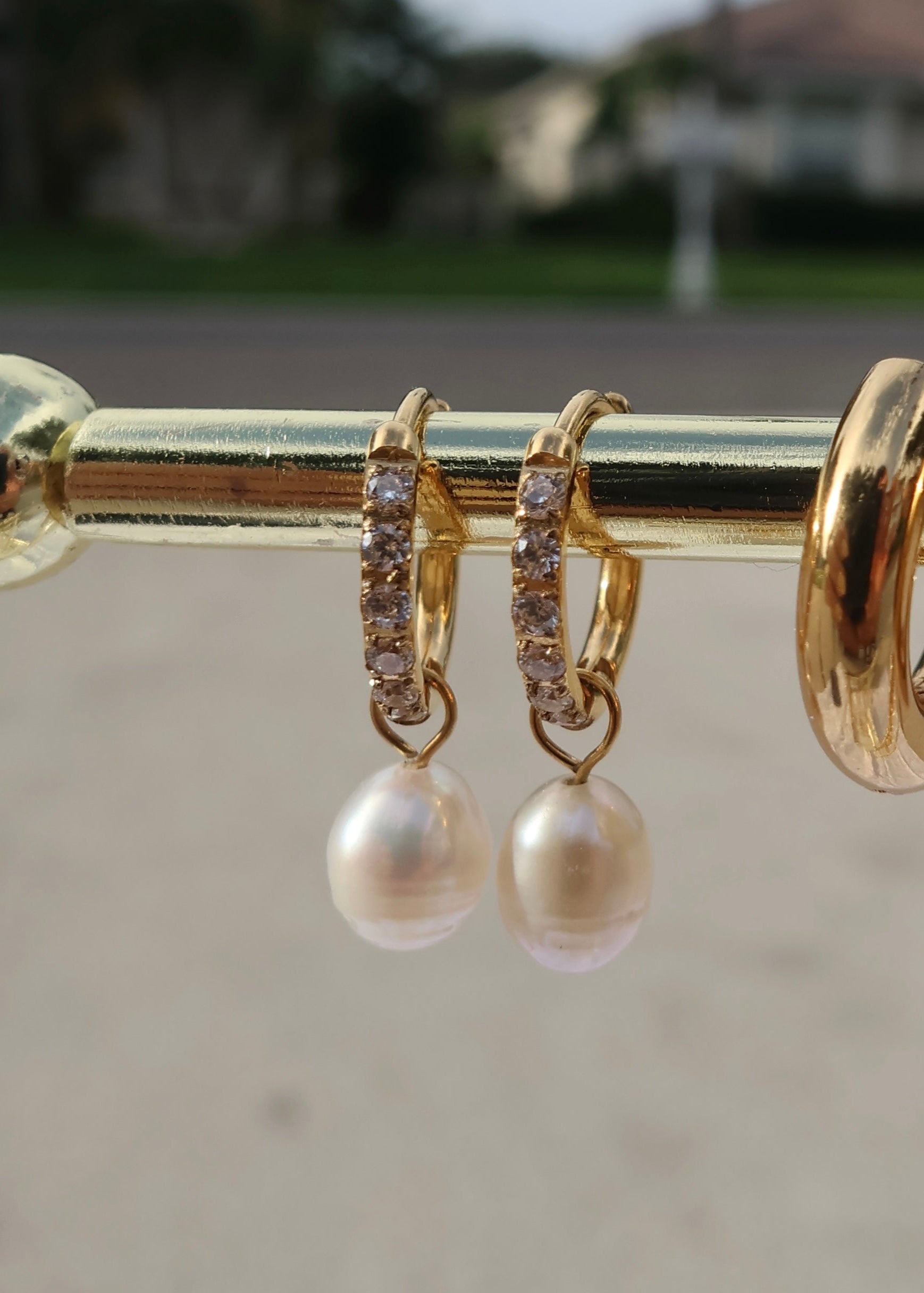 Gold hoop earrings with pearl drops on a gold bar against a blurred outdoor background